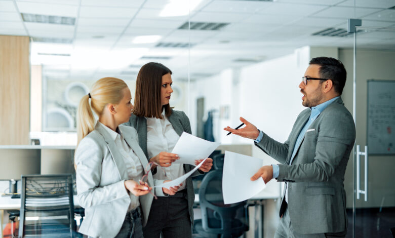 Business people arguing in modern office, holding paper documents. Two businesswomen surprised with annoyed businessman.